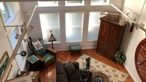 Ein geräumiges Wohnzimmer mit einer großen Couch, einem Couchtisch und einer Holzkabine. Mehrere Fenster mit Jalousien, eine Lampe und eine kleine Bank sind vorhanden.