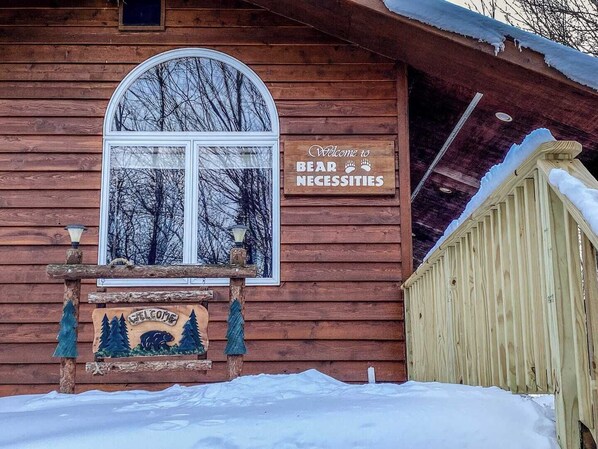 Una cabaña de madera con un cartel que dice 'Welcome to Bear Necessities', una ventana con vistas a árboles desnudos y un terreno cubierto de nieve.