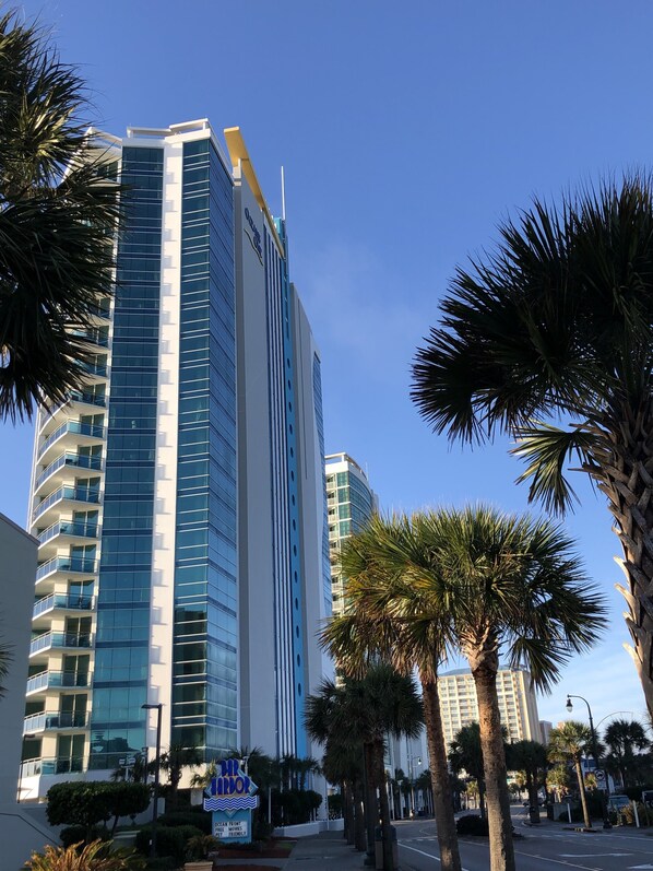 A tall, modern hotel with a glass facade and multiple balconies, surrounded by palm trees and a clear blue sky.