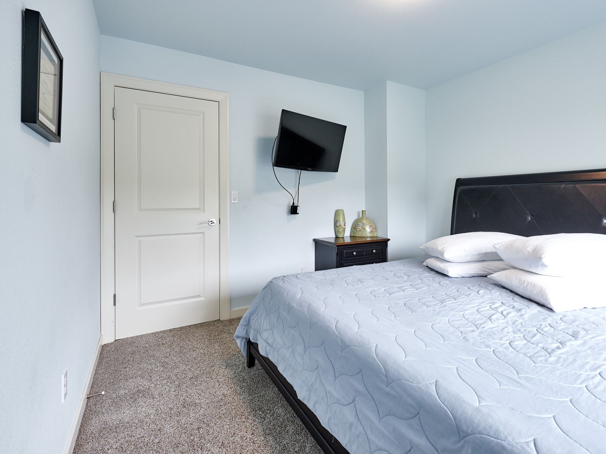A bedroom with a bed, a TV mounted on the wall, a door, and a small nightstand with decorative items.