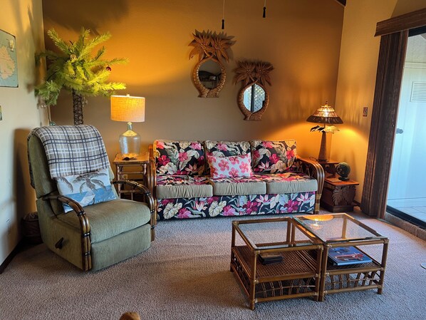 A cozy living room with a green armchair, a floral sofa, a glass coffee table, and two wall mirrors.