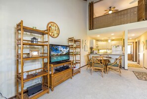 Een woonkamer met een televisie, houten schappen, een eethoek met rieten stoelen en een keuken op de achtergrond.