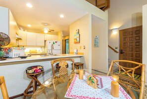 A modern kitchen with white cabinets, a microwave, and a refrigerator. There is a dining area with a glass table, wooden chairs, and a small island with a fan.