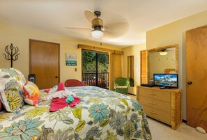 A bedroom with a bed, a ceiling fan, a dresser, a television, and a window with a view of greenery.