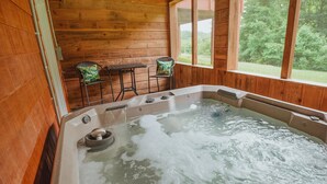 A wooden cabin with a hot tub, two chairs with floral cushions, and a window overlooking greenery. - Log Cabin, Hot Tub, Fire Pit, Near Yellowwood (Nashville)
