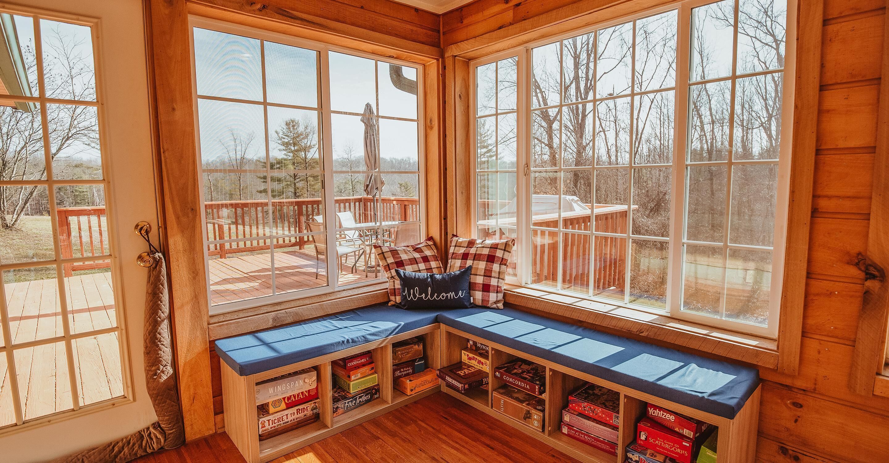 A cozy cabin with a built-in bench and shelves filled with board games, large windows offering a view of the deck and surrounding trees.