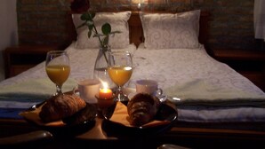 En pænt redt seng med et gråt og hvidt mønstret sengetæppe, en vase med en enkelt rose og en bakke med croissanter og drikkevarer på et træbord.