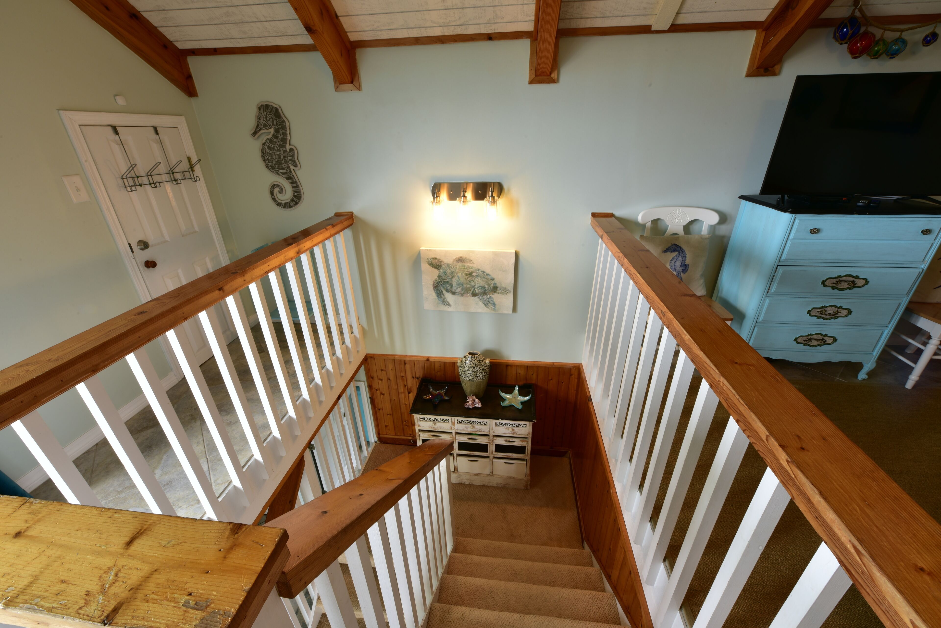 A wooden staircase with white railings leading to a landing with a wall-mounted light fixture, a seahorse decoration, and a turtle painting.
