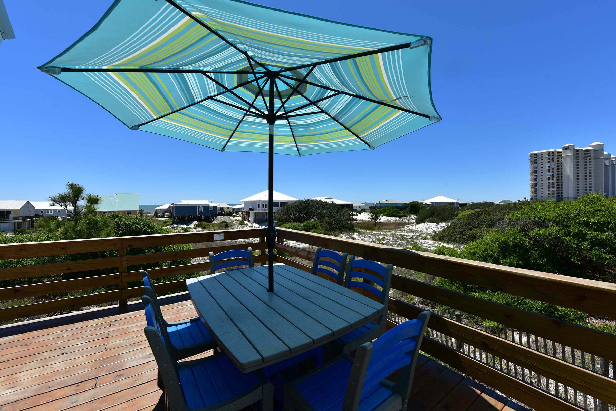 A wooden deck with a table and chairs, a striped umbrella, and a view of the beach and buildings.