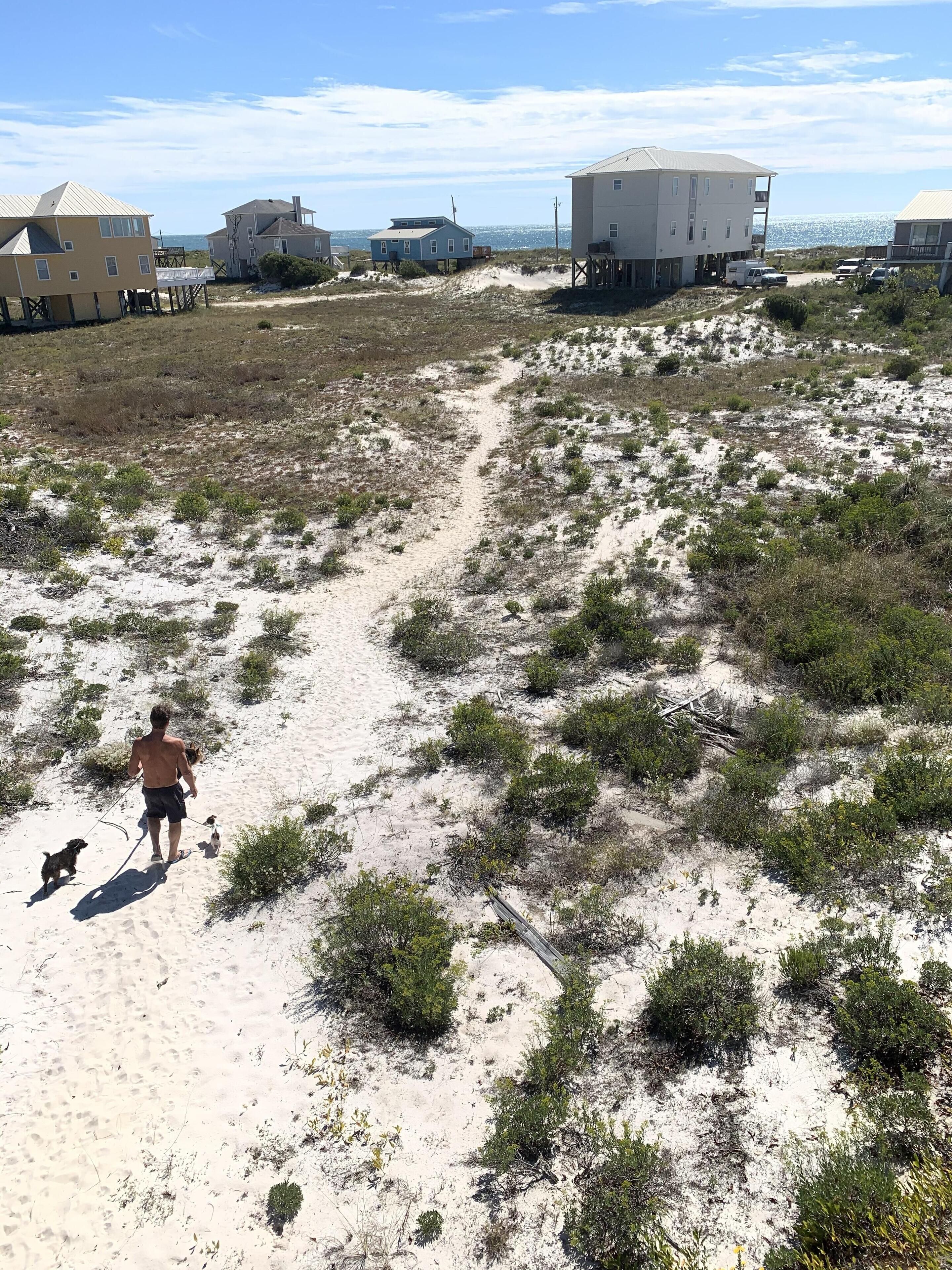 A sandy beach area with sparse vegetation and several buildings in the background.
