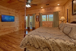 A wooden cabin bedroom with a bed, a ceiling fan, a TV, and a window with curtains.