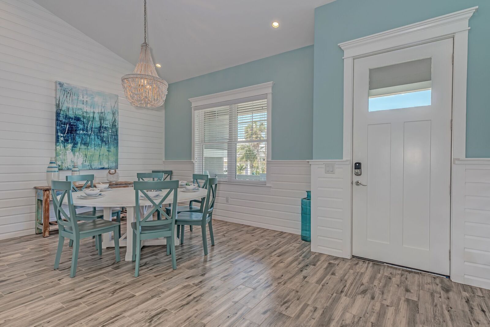 Un coin repas avec une table blanche, des chaises de couleur turquoise et un lustre. Une grande fenêtre avec des stores et une porte dotée d’une petite fenêtre sont également présents.