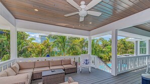 A covered outdoor area with a wooden ceiling, a white ceiling fan, a beige sofa, wicker furniture, and a view of a swimming pool and tropical vegetation.