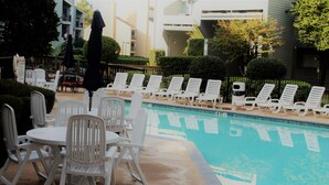 Un espace aménagé au bord de la piscine, avec des chaises longues et des tables blanches, une piscine, et un bâtiment en arrière-plan.