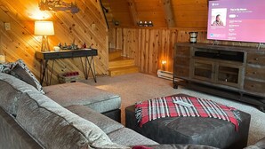 En mysig stuga med träpanel på väggarna, en hörnsoffa, en öppen spis och en tv som visar en musikspelare.