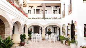 Un patio con sillas y mesas blancas, plantas en macetas y flores rojas.