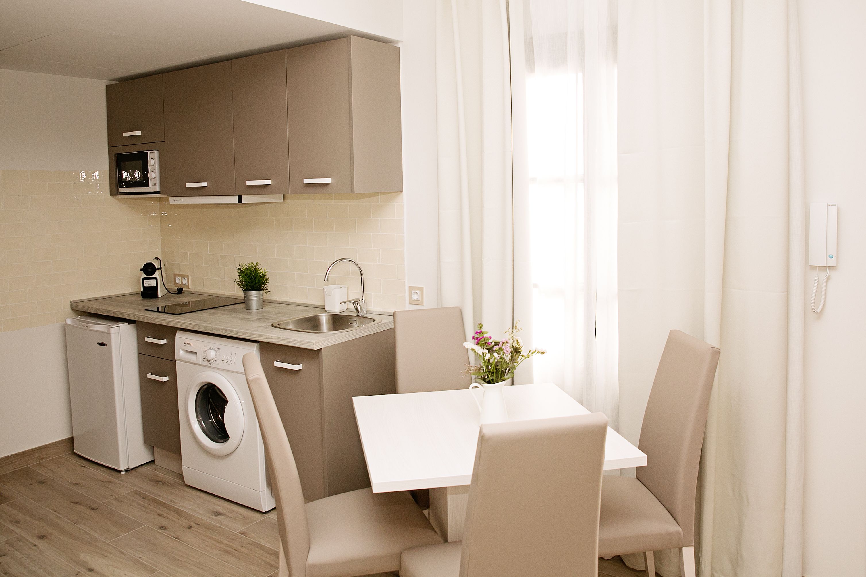 A compact kitchen with a washing machine, a microwave, and a sink. There is a small table with chairs and a vase with flowers.