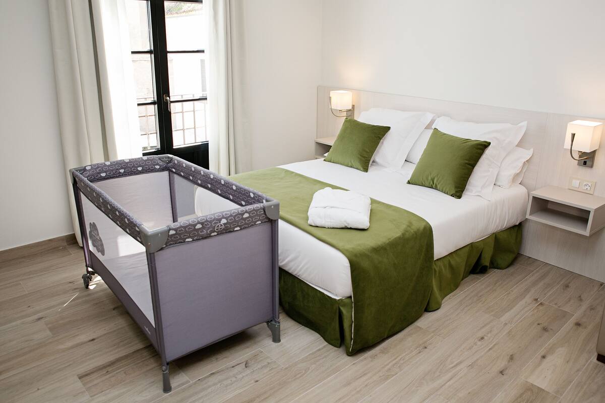 Une chambre d’hôtel avec un lit, un berceau, une table de chevet et une fenêtre avec des rideaux.