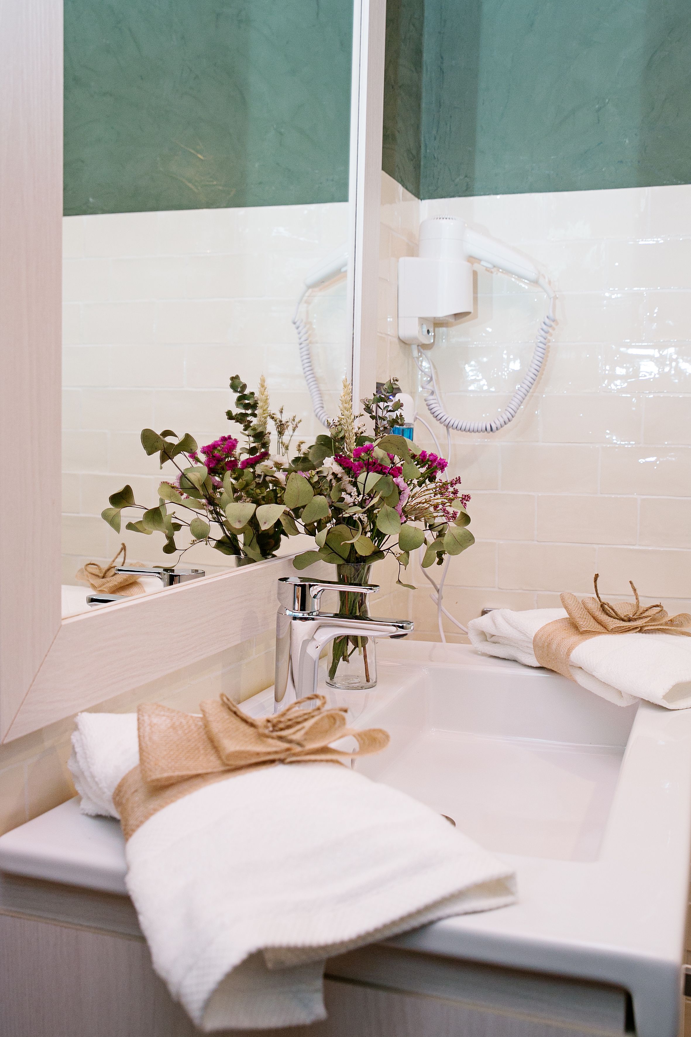 A bathroom with a mirror, a sink, and a shower. There is a towel rack with towels and a vase with flowers on the sink.