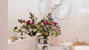 A bathroom with a mirror, a sink, and a shower. There is a towel rack with towels and a vase with flowers on the sink.
