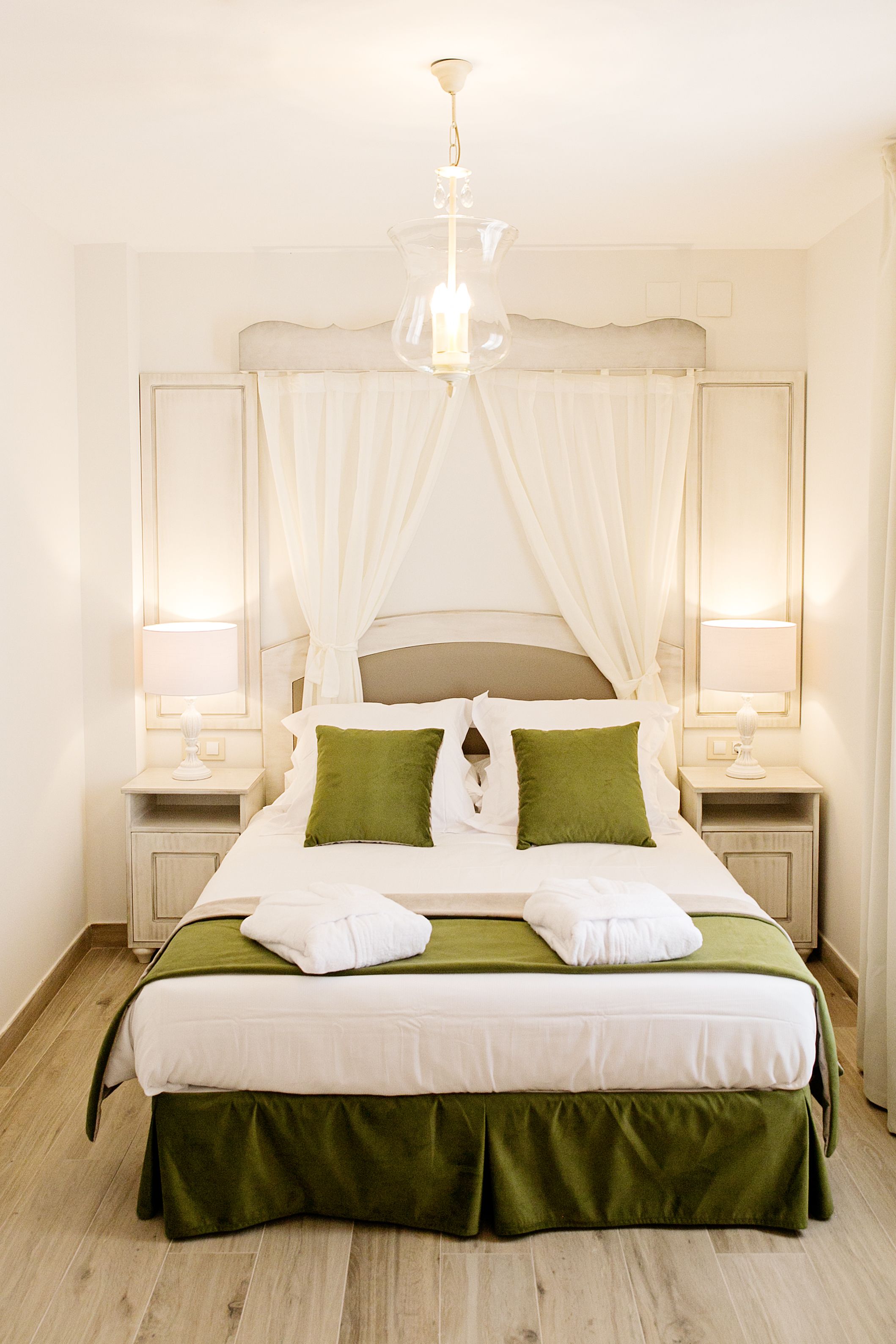 A neatly made bed with white and green bedding, two bedside lamps, and a chandelier above.