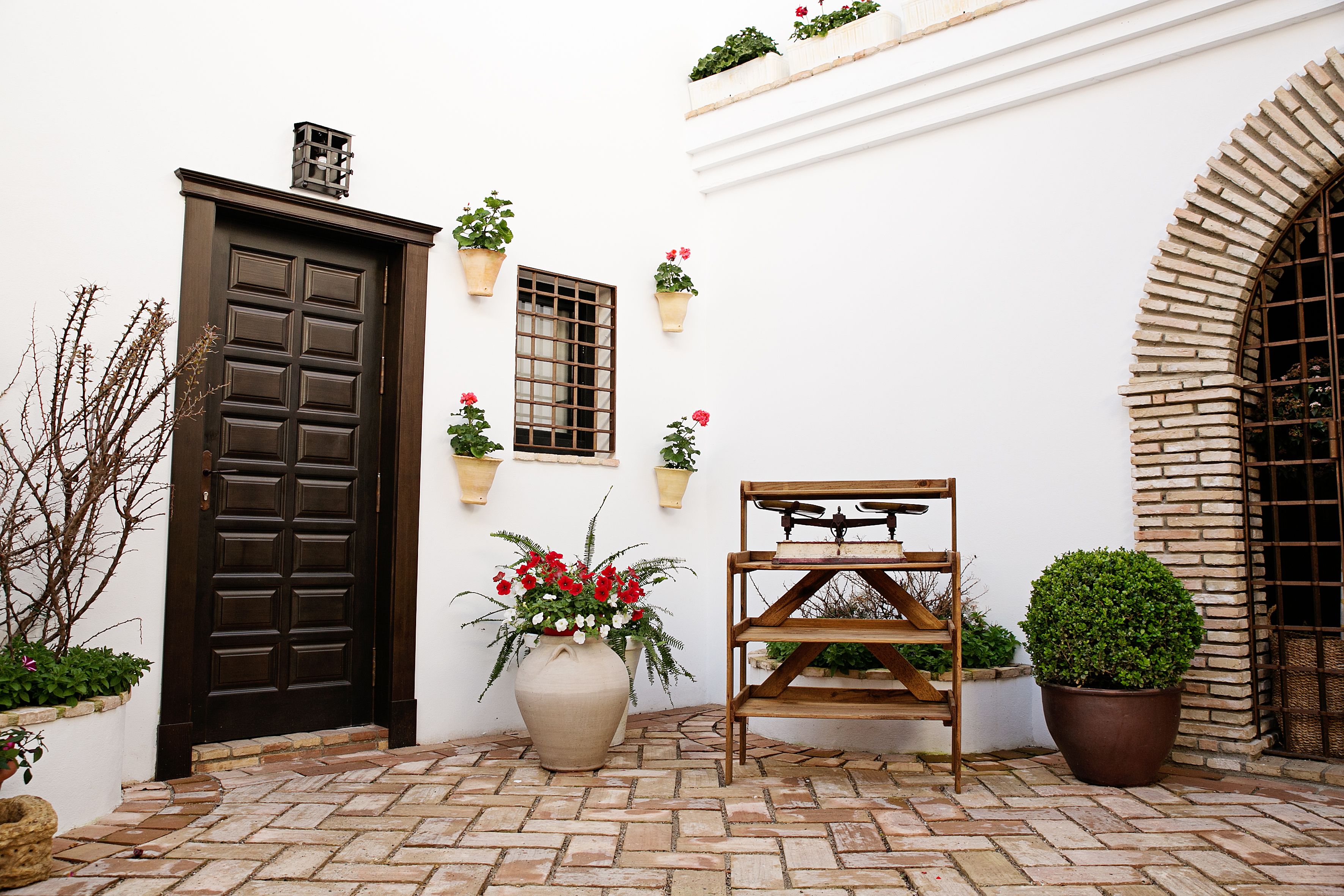 A courtyard with a brown door, potted plants, a wooden shelf, and a brick floor.
