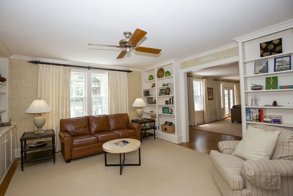 A living room with a brown leather sofa, a round coffee table, a white armchair, and a bookshelf filled with books and decorative items.