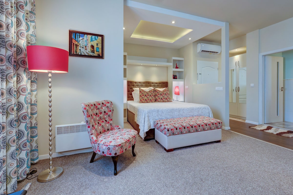 A bedroom with a bed, a chair, a bedside table, a lamp, and a picture on the wall.