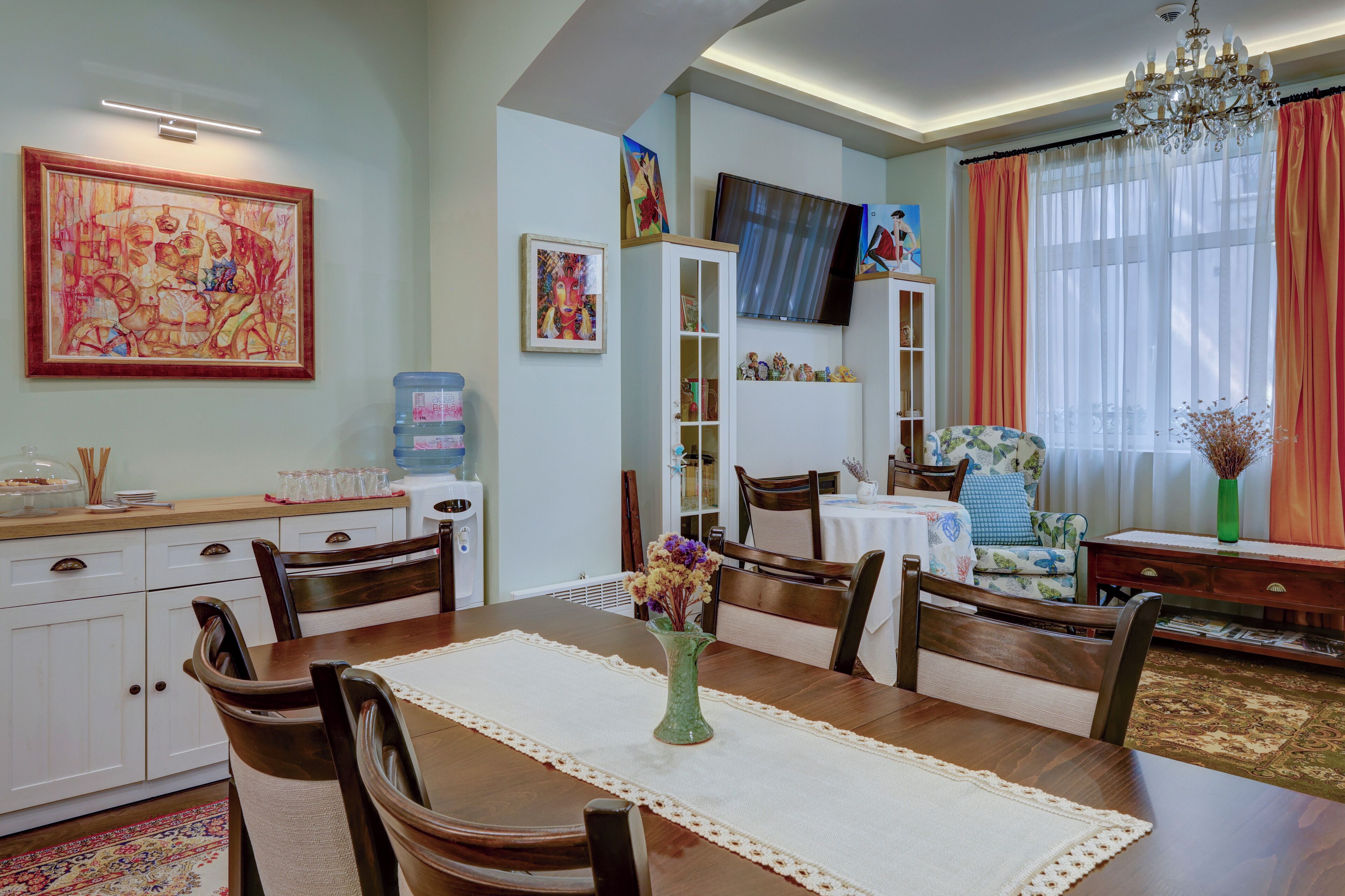 Un comedor con una mesa de madera, sillas y un jarrón de flores. Hay un televisor montado en la pared y un dispensador de agua en la encimera.