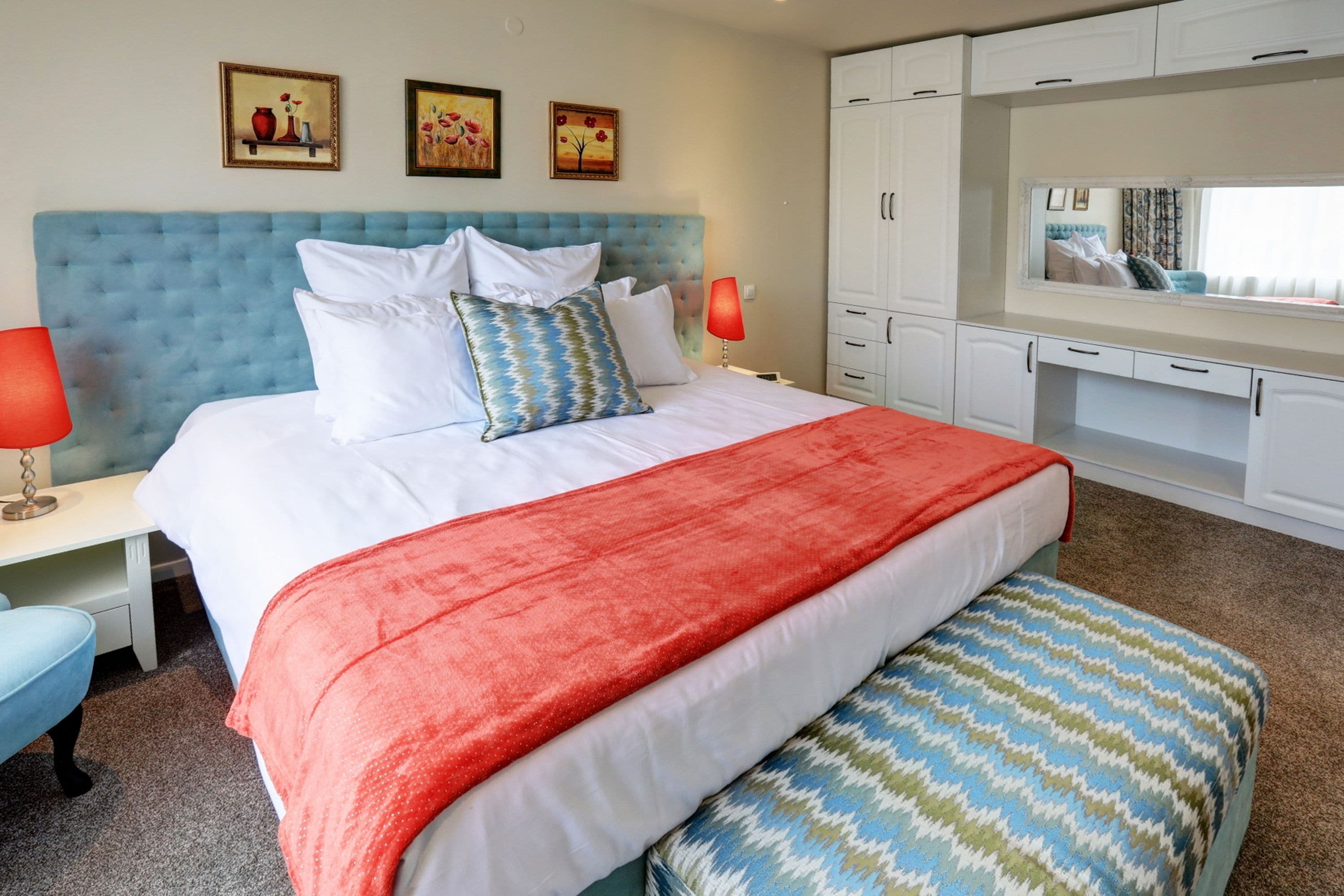 Un dormitorio con una cama grande, ropa de cama blanca, una manta roja y un cabecero azul. Hay tres cuadros en la pared, una mesita de noche con una lámpara y un armario con espejo.
