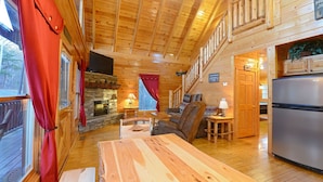 A cozy cabin interior with a stone fireplace, wooden furniture, a dining table, and a kitchen area. - Charming Cabin with Private Hot Tub & Pool Table (Sevierville)