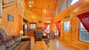 A log cabin interior with a living area, dining table, kitchen, and a large window with red curtains. - Charming Cabin with Private Hot Tub & Pool Table (Sevierville)