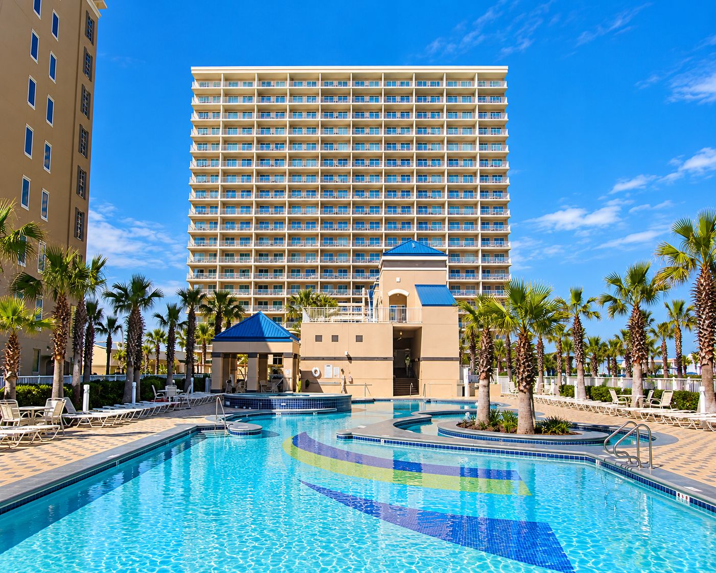 A hotel with a large pool area, palm trees, and a multi-story building in the background.