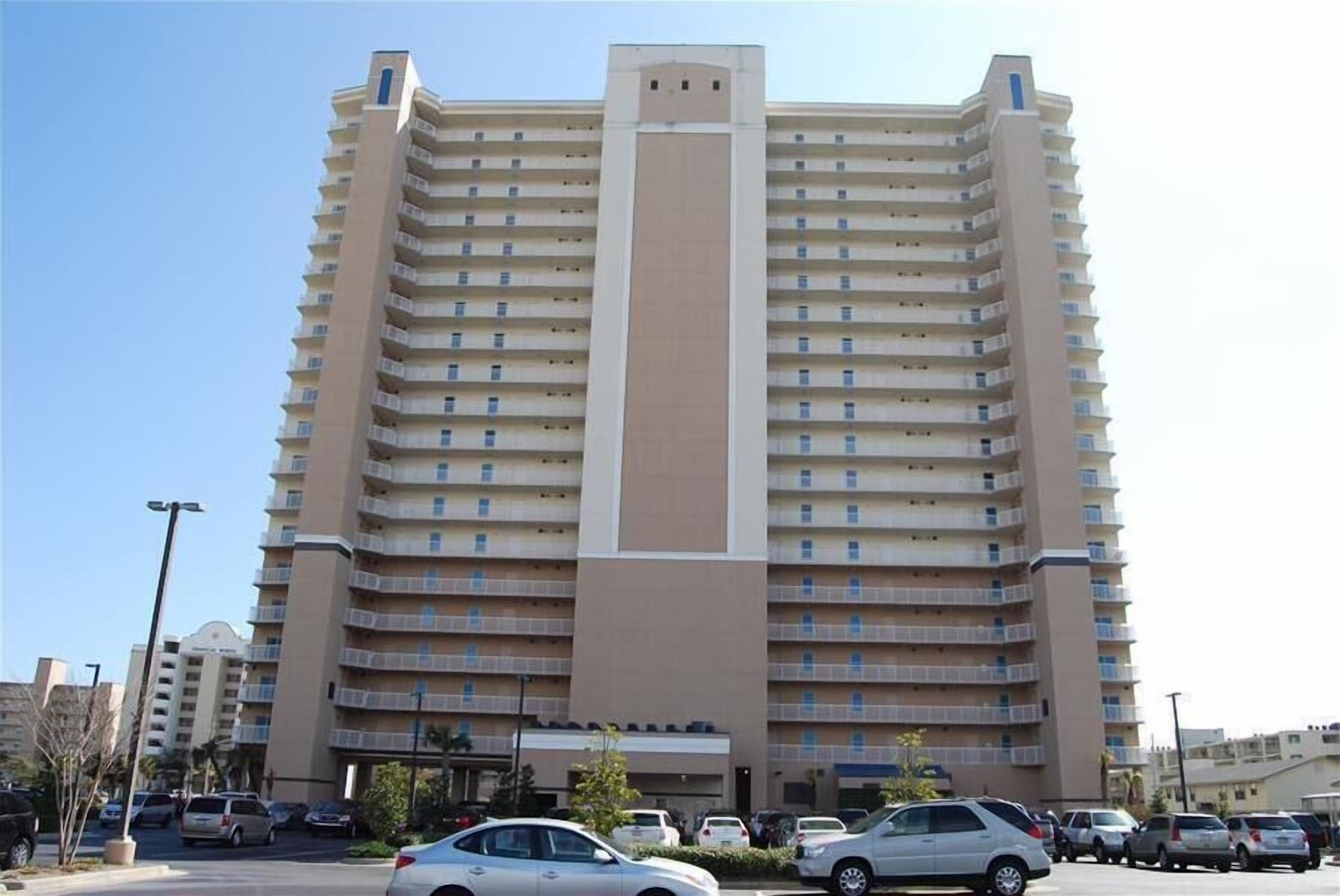 A multi-story hotel with a central vertical feature, balconies on each floor, and a parking lot in the foreground.