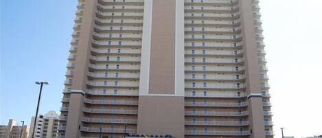 A multi-story hotel with a central vertical feature, balconies on each floor, and a parking lot in the foreground.