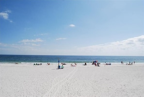 Une vaste plage de sable où l’on peut voir des gens se prélasser au soleil et se baigner, un ciel bleu dégagé et l’océan en toile de fond.