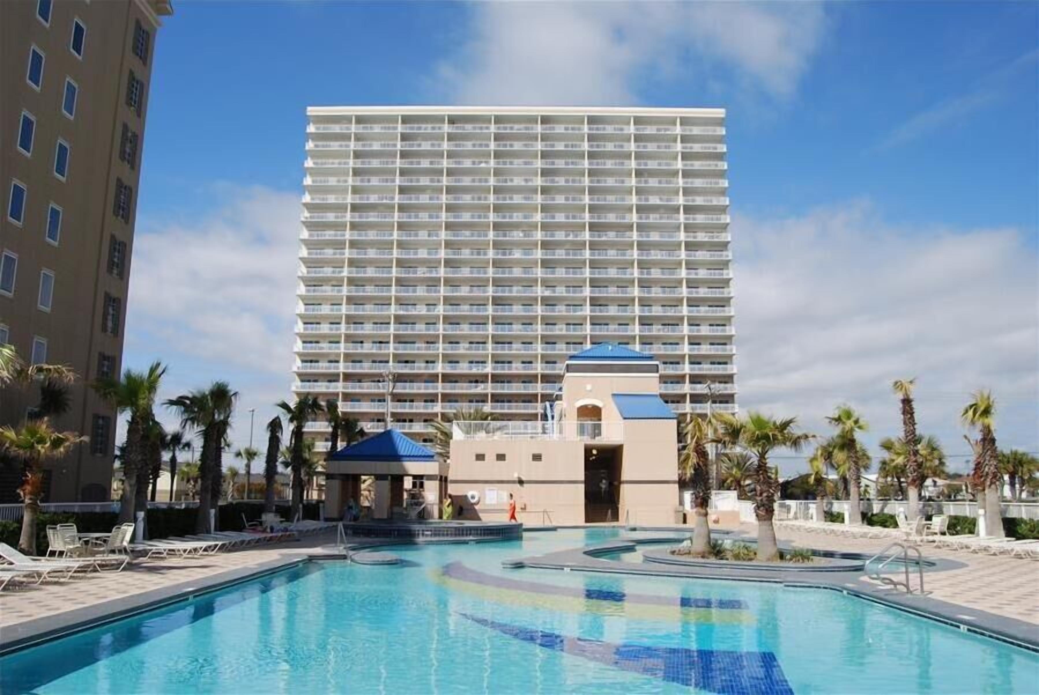 Un hotel con un amplio espacio de piscina, palmeras y un edificio de varios pisos al fondo.