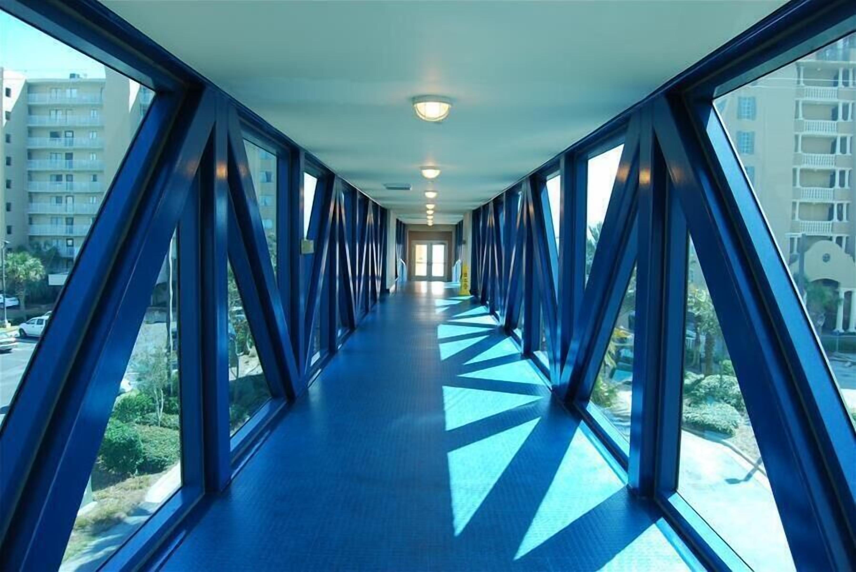 A corridor with a blue floor and large windows on both sides, leading to a door at the end.