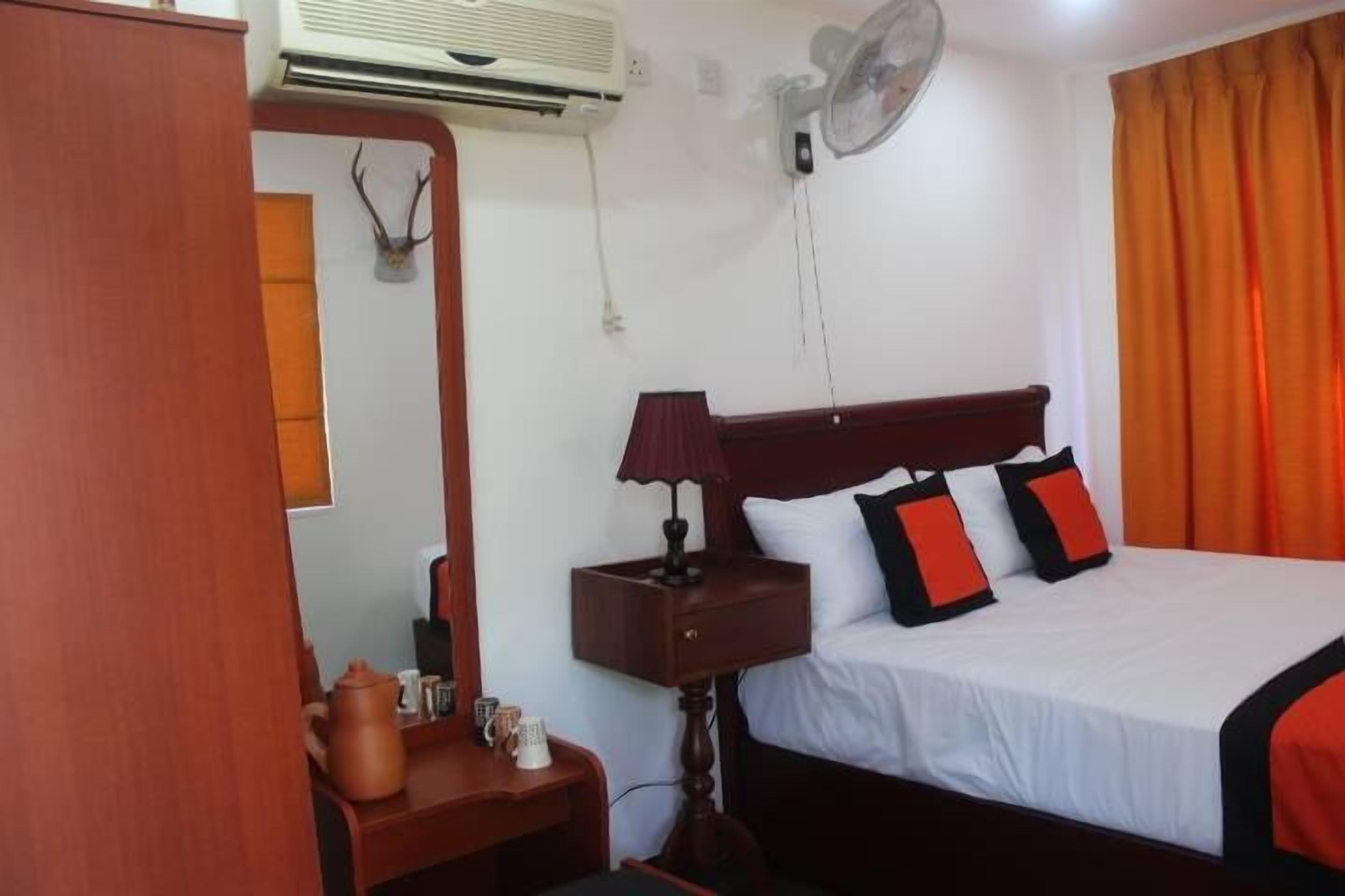 Habitación de hotel con cama, mesita de noche, lámpara, ventilador, aire acondicionado, espejo y ventana con cortinas.