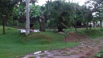 A park with a small wooden bench, a large decorative wheel, and various trees including palm trees.