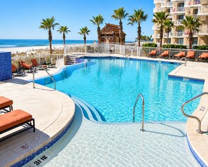 A swimming pool with a curved edge, surrounded by lounge chairs and palm trees, with a beach and ocean in the background. - Spacious Gulf-Front 4BR Condo | Sleeps 10 | Huge Balcony | Indoor & Outdoor Pools (Orange Beach)
