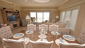 A dining area with a wooden table set for six, surrounded by six chairs. There is a television and a seating area with a couch and chairs in the background. - Relax and Catch a couple of fish! 🎣 ~ There is something for everyone! (Orange Beach)