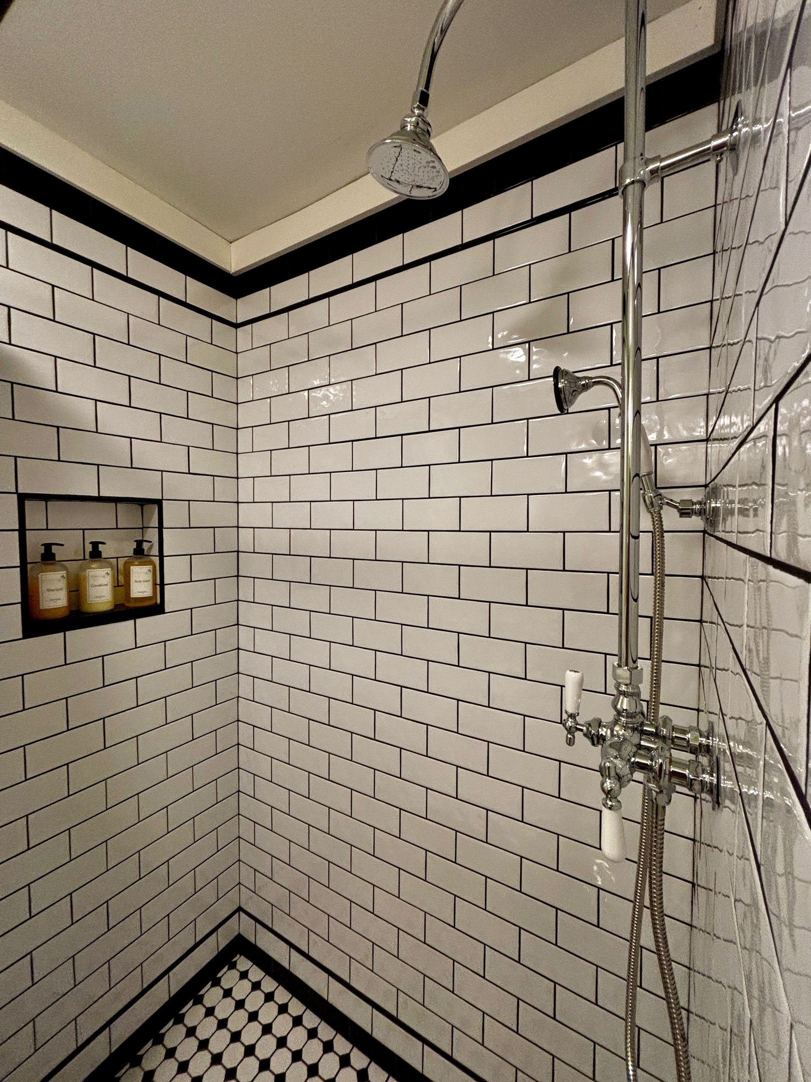 A bathroom with white subway tiles, a shower with a handheld showerhead, and a shelf with three bottles.