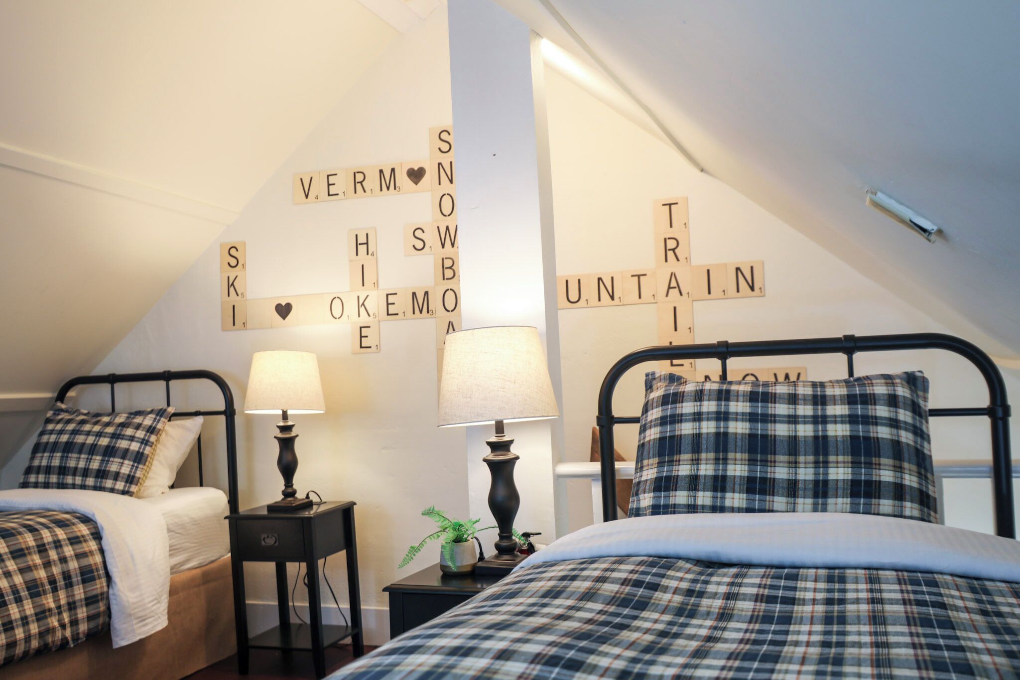 A cozy bedroom with two beds, each with plaid bedding and a lamp. The walls have words spelled out in tiles, and there's a small plant on the nightstand.