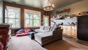 A spacious living room with a grey sectional sofa, a coffee table, and an American flag on the floor. There are large windows providing natural light and a kitchen area with wooden cabinets and a microwave.