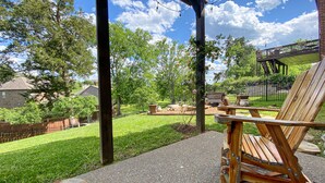 A covered patio with a wooden chair, a concrete path, and a view of a backyard with a lawn, trees, and a wooden deck.