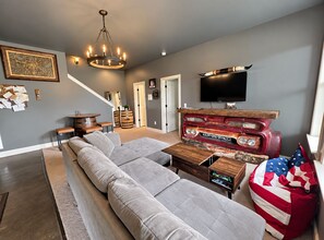 A modern living room with a sectional sofa, a vintage car display, a television, and a chandelier. - Indulge in Relaxation:King Bed Suite with Park Access for Your Ultimate Getaway! (Nashville)