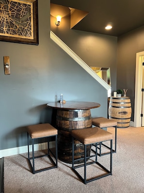 A room with a round wooden barrel table, two cushioned stools, a wall-mounted light fixture, a wooden barrel, and a staircase. - Indulge in Relaxation:King Bed Suite with Park Access for Your Ultimate Getaway! (Nashville)