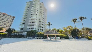 Ein Hochhaus-Hotel mit mehreren Balkonen, ein Sandstrand mit Liegestühlen und Palmen.