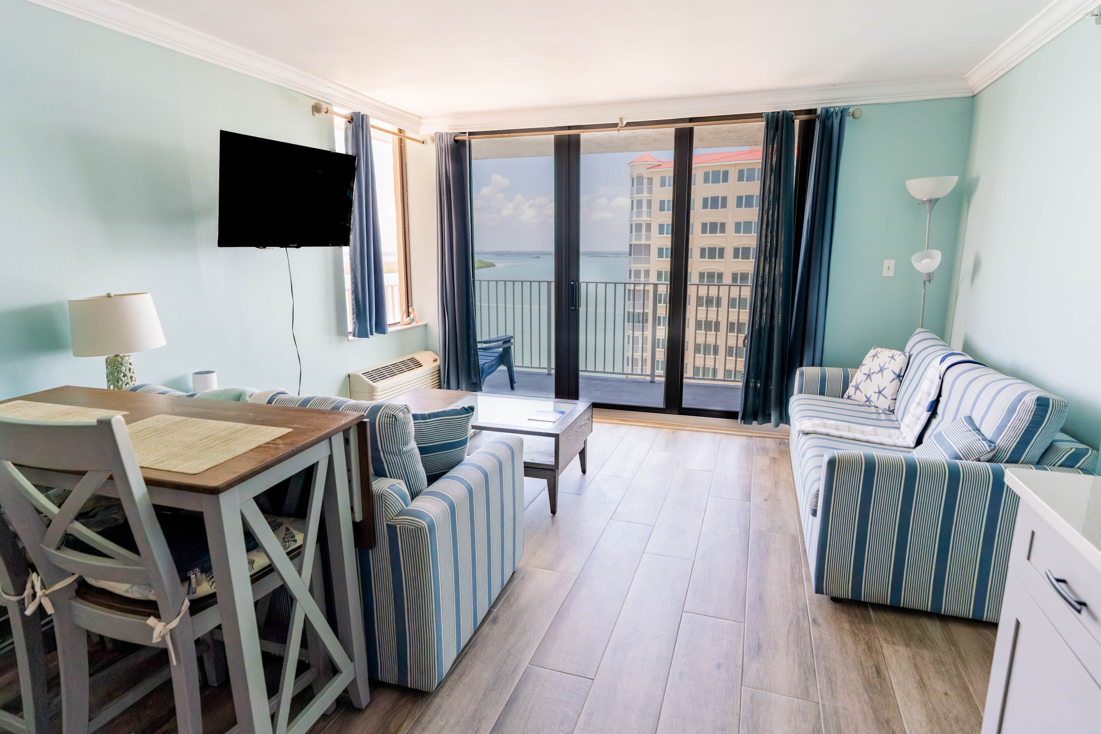 A modern living room with a dining area, a flat-screen TV mounted on the wall, and a view of a building across the balcony.
